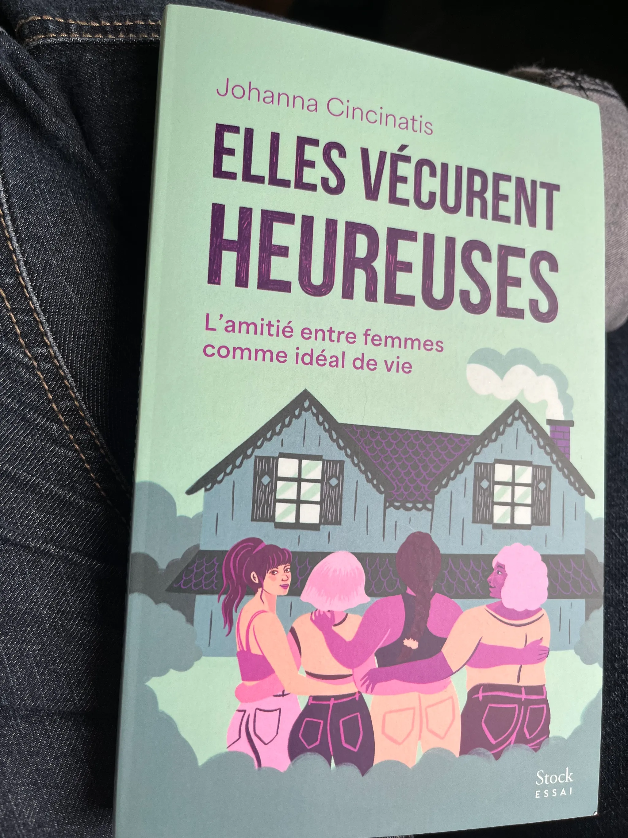 Le livre titré Elles vécurent heureuses, posé sur ma jambe gauche, j'ai un jean bleu. La couverture et bleu-verte, avec quatre femmes dessinées devant une maison. Le sous-titre est : L'Amitié entre femmes comme idéal de vie.