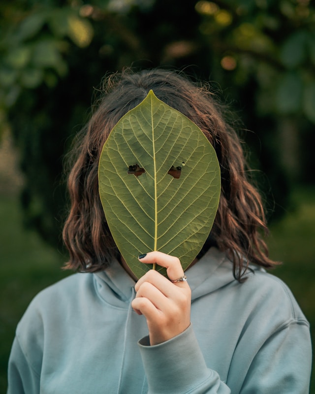 Une femme cachée derrière une feuille d'arbre, avec uniquement deux trous pour les yeux qui laissent apparaître son regard